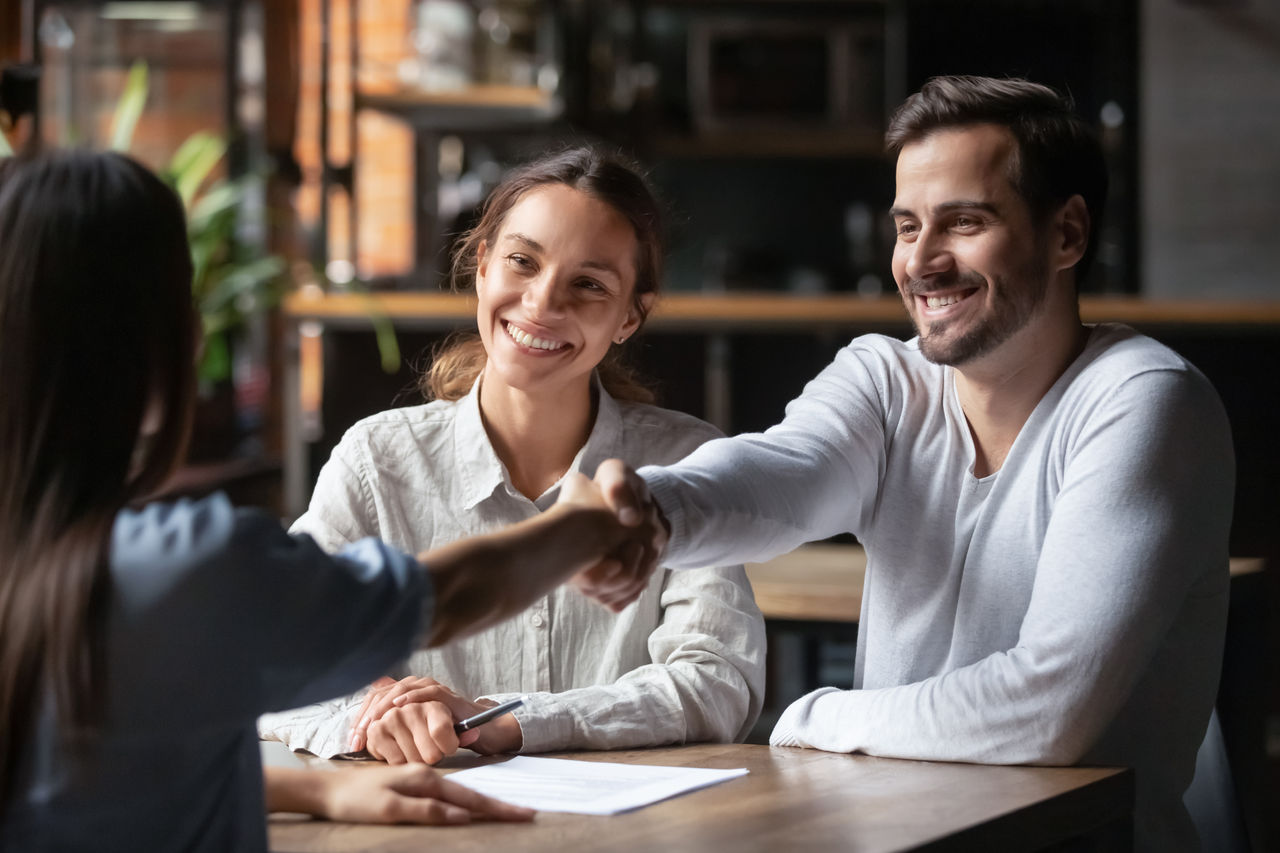 Happy smiling millennial couple handshake get acquainted with female real estate agent meeting together in cafe, excited husband and wife shake hand of broker or banker thanking for help